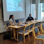 Pöydän takana istumassa kaksi naista, toisella edessään tietokone. Taustalla valkokangas, missä sinisellä pohjalla tekstiä ja numero 72.