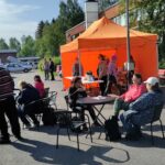 Torilla. Taustalla oranssi telttakatos, muutama ihminen sen sisällä seisomassa. Edessä pyöreitä pöytiä pari, niissä ihmisiä istumassa kahvia juomassa.