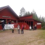 Taustalla punamultaisia rakennuksia; katettu ulkoilmateatteri. Edessä seisomassa kolme ihmistä.