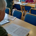 Pöytien välissä seisomassa nainen vihko kädessään. Etualalla mies istumassa, hänellä edessään tehtävä sivuja.