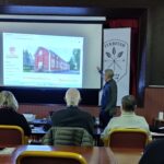 Taustalla valkokangas, missä talonkuva. Oikealla edessä mies seisomassa, edessä selin ihmisiä istumassa.