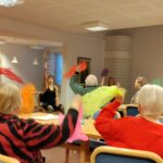 Ihmisiä istumassa pöydissa. He huiskuttavat värikkäitä huiveja. Ohjaajat istuvat edessä.