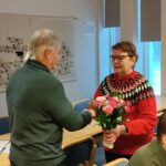 Mies seisomassa selin. Edessään nainen. He kättelevät. Naisella punainen kukkakimppu kädessään.