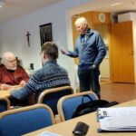 Mies seisomassa paperi kädessään. Etualalla kaksi miestä istumassa pöydän äärellä, toinen selin.