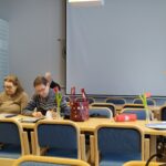 Oikealla nainen, punaisessa puserossa seisomassa, paperi kädessään. Istumassa pöydissä vasemmalla kolme henkilöä.