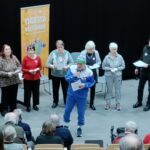 Edessä seisomassa puoliympyrässä 9 ihmistä. Yksi ihminen etualalla. Taustalla vasemmalla flyygeli, jonka edesä istumassa nainen. Yleisö istumassa selin katsojaan päin.