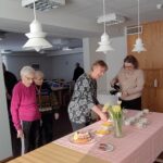 Pöydällä kahvikuppeja, tiikerikakku, keltaisia tulppaaneja. Pöydän takana jonottamassa ihmisiä. Pöydän päässä nainen kaatamassa lasikannusta kahvia kuppiin.