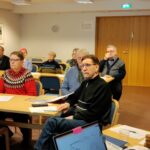 Pöydissä istumassa ihmisiä, katsovat eteenpäin. Etualalla vinoittain olevassa pöydässä istumassa selin mikkiä pitelevä nainen.